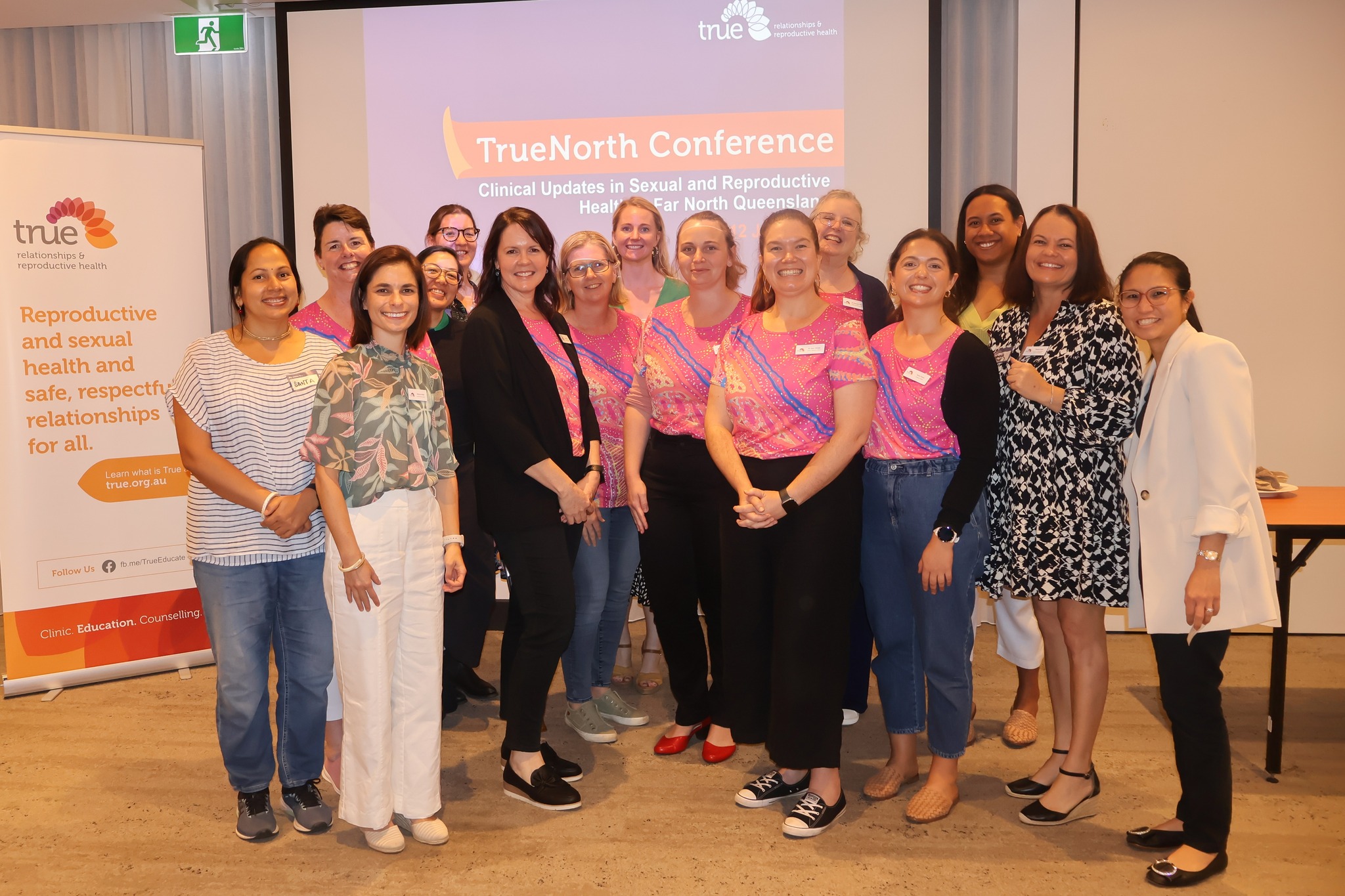A group of women, mostly in pink t-shirts stand in front of a sign that says 'True North Conference'. A group of women, mostly in pink t-shirts stand in front of a sign that says 'True North Conference'.